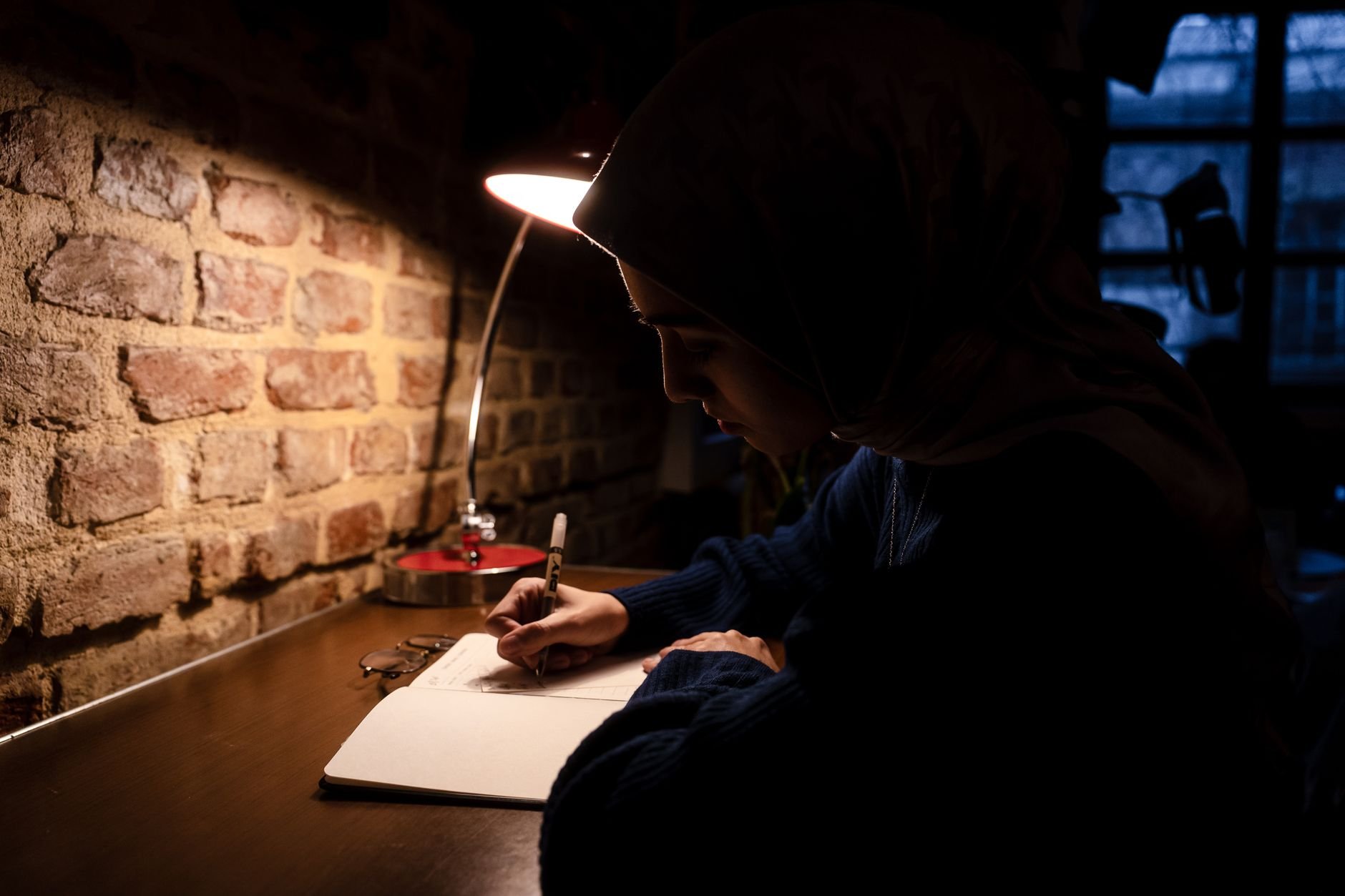 A young woman wearing a hijab writes in a notebook under warm lamp light